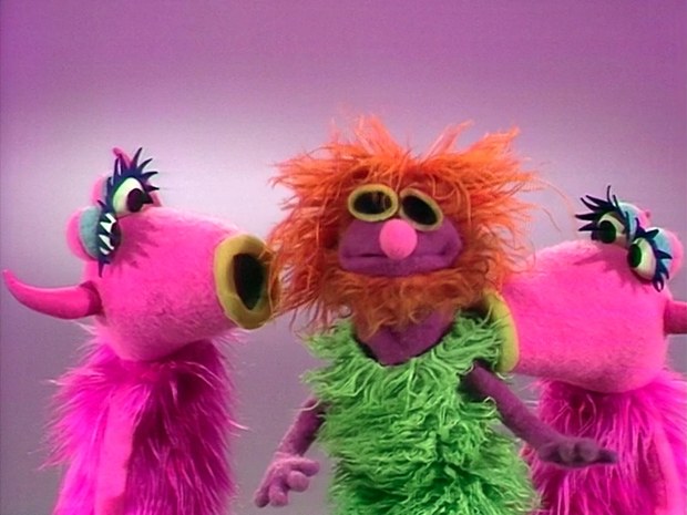Three furry puppets: two pink ones and an orange-haired green one.